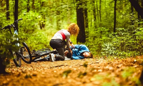 Young sportswoman leaning over male cyclist with broken leg on forest path and trying to understand what to do